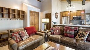 A cozy living room with leather sofas, a wooden coffee table, and a kitchen area with wooden cabinets and modern appliances. - Phoenix 108 4 Bedroom Townhouse by RedAwning (Steamboat Springs)