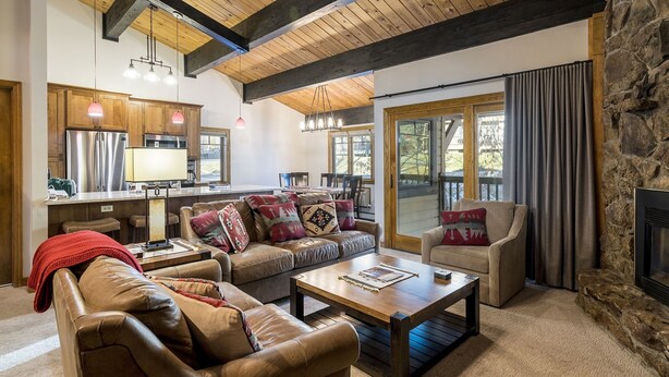 A spacious living room with a leather sofa, a coffee table, and a fireplace. There is a kitchen area with wooden cabinets and a large window. - Phoenix 108 4 Bedroom Townhouse by RedAwning (Steamboat Springs)