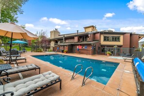 A swimming pool with lounge chairs and umbrellas, a building with a sign, and a fenced area with greenery. - Phoenix 108 4 Bedroom Townhouse by RedAwning (Steamboat Springs)
