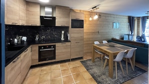 A modern kitchen with wooden cabinets, a black countertop, and a dining area with a table and chairs. - Apartment for 4 people on the slopes (Lanslevillard)