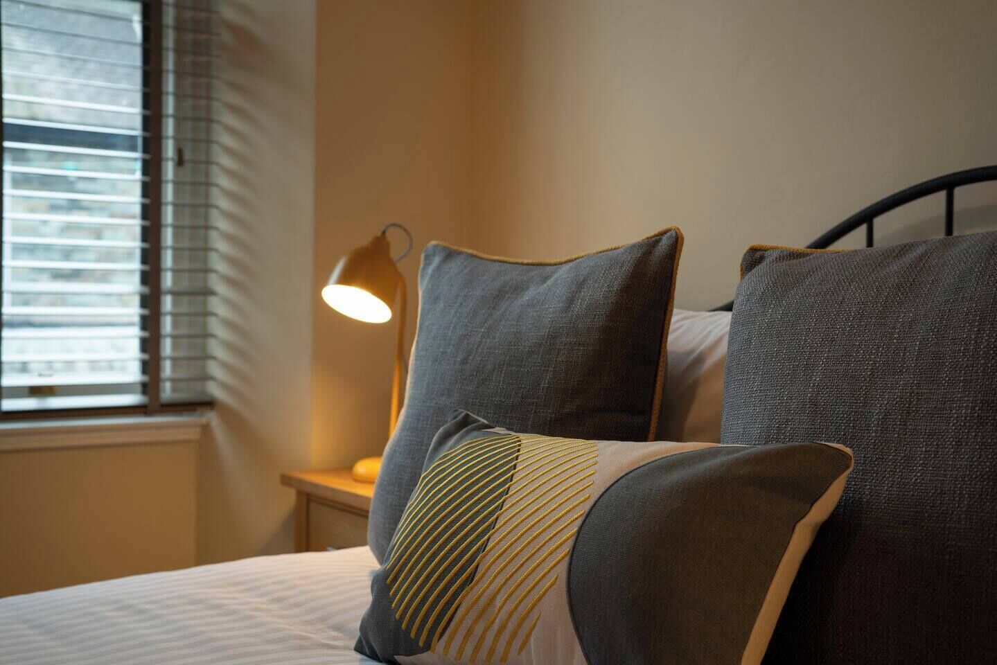 A neatly made bed with grey and striped pillows, a bedside lamp, and a window with blinds.
