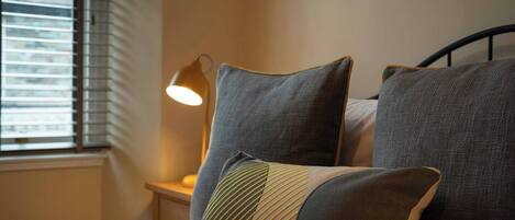 A neatly made bed with grey and striped pillows, a bedside lamp, and a window with blinds.