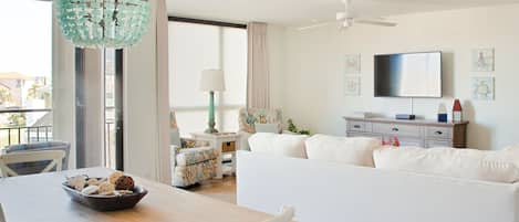A modern living room with a white sofa, a dining table with a bowl of snacks, a chandelier, and a television on the wall.