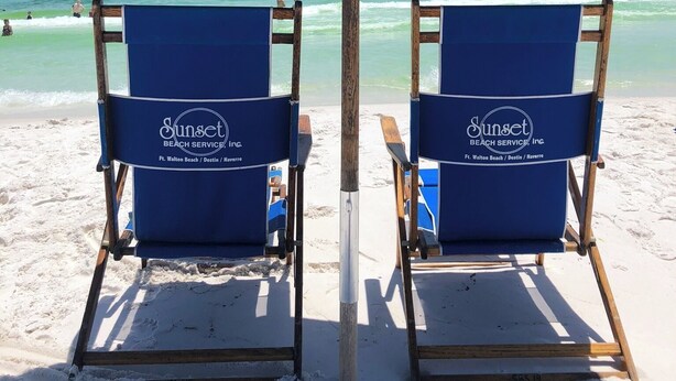 Dos sillas de playa bajo una sombrilla azul, con el texto "Sunset Beach Service Inc." en los respaldos.