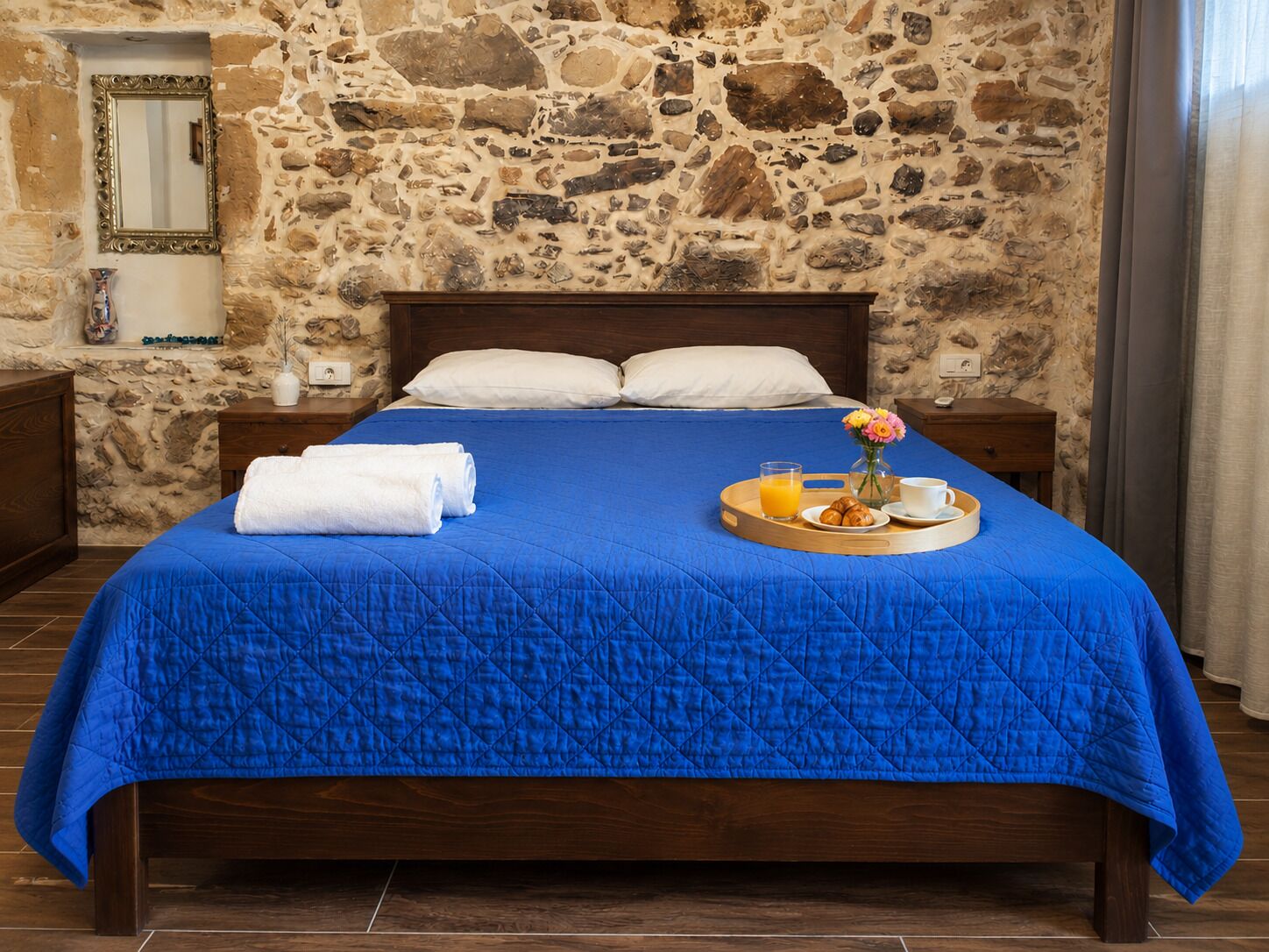 Un lit avec une couette bleue, des oreillers blancs, une table de chevet en bois, un mur en pierre, un miroir et un plateau avec des croissants, du jus et une tasse.