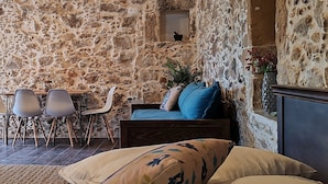 Una habitación con pared de piedra, una cama con almohadas color beige y azul, una pequeña mesa con sillas y una planta.