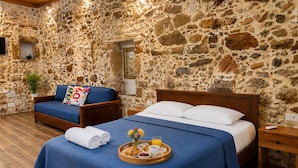 Una habitación con pared de piedra, cama con colcha azul, banco de madera con cojín y bandeja con alimentos para el desayuno.
