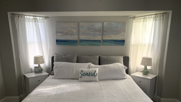 Une chambre à coucher avec un grand lit, deux tables de chevet avec des lampes, une tête de lit ornée d’un tableau et des rideaux légers.
