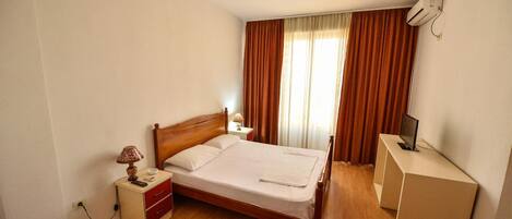 A hotel room with a wooden bed, a desk with a computer, a small bedside table with a lamp, and a red bench.