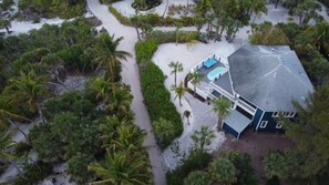 Vue aérienne d’une maison dotée d’une piscine, entourée d’une végétation tropicale luxuriante et d’une zone sablonneuse.