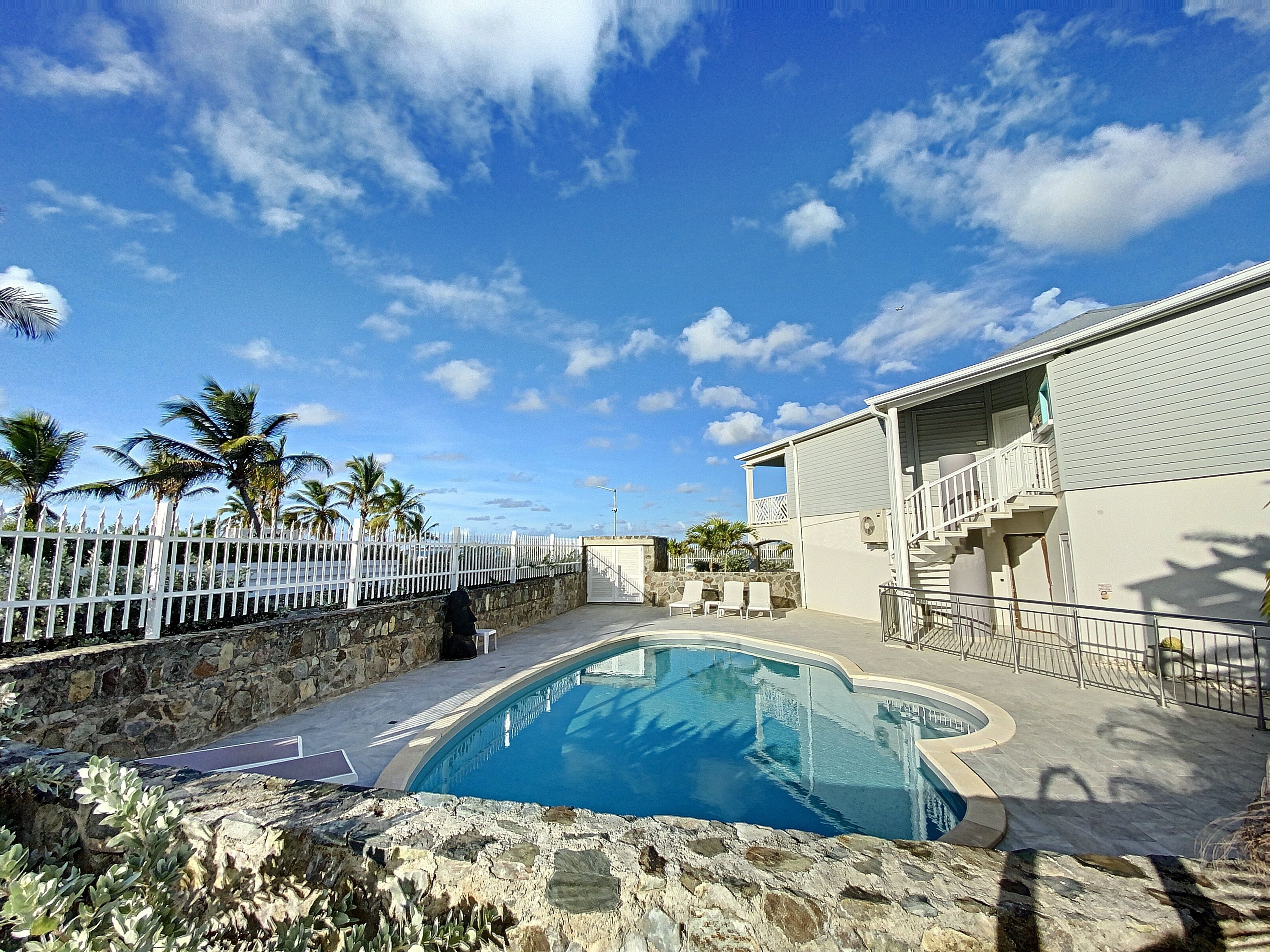 A pool area with a stone wall, palm trees, and a building with a balcony.