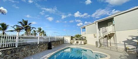 A pool area with a stone wall, palm trees, and a building with a balcony.