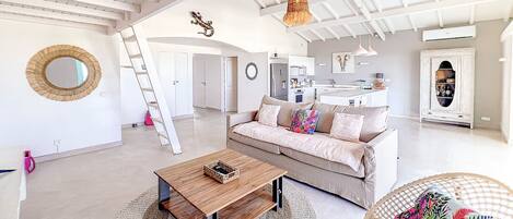 A spacious living area with a high ceiling, a wooden coffee table, a sofa with colorful pillows, and a wicker chair.