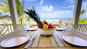 A table with a fruit bowl, plates, and cutlery, overlooking a beach with palm trees and a clear sky. - Princess Manon, Beachfront, Feet in water,  Ocean view, Pool (Saint-Martin)