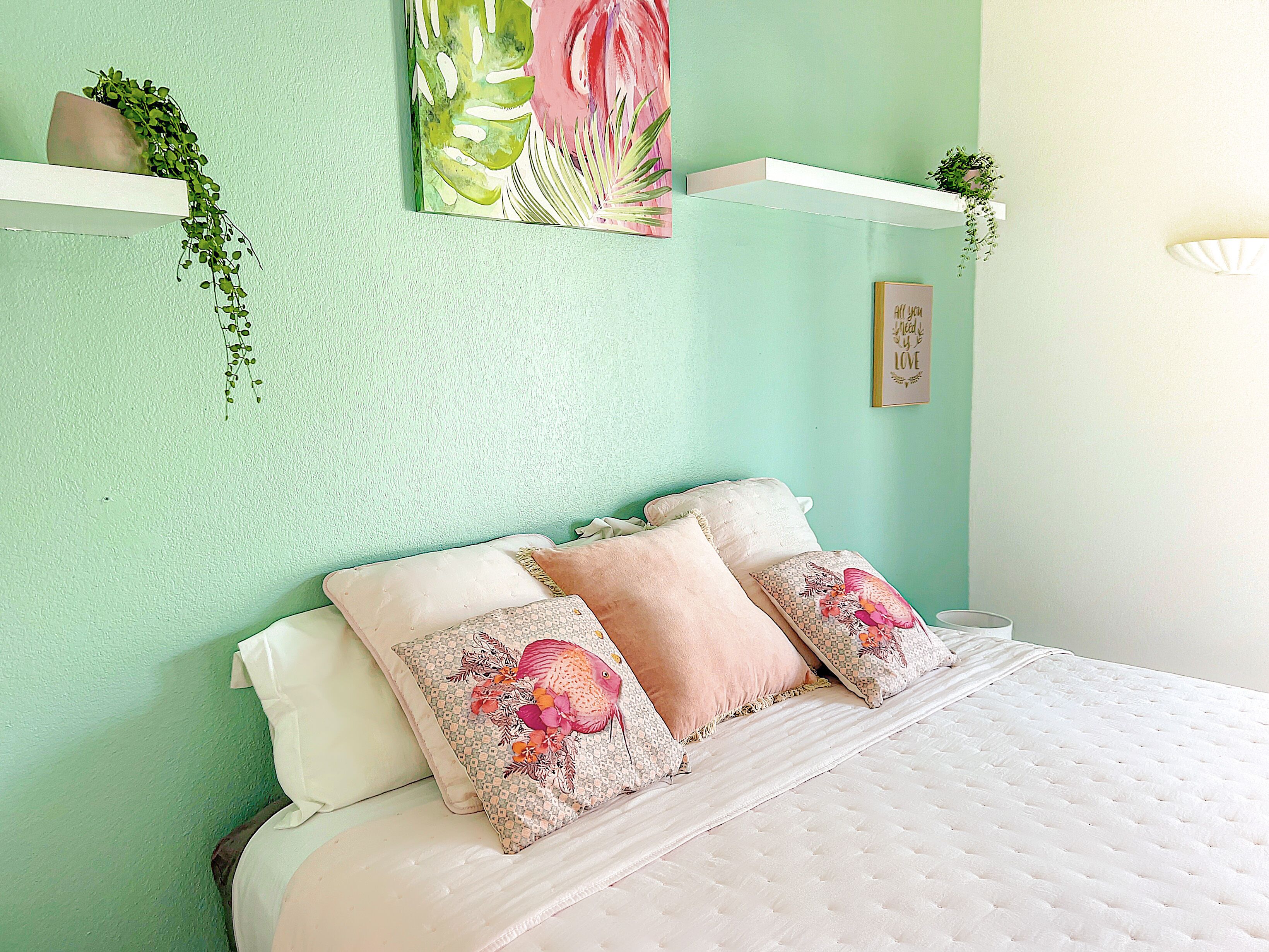 Un dormitorio con cama, almohadas, un estante en la pared con plantas y un cuadro.