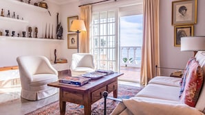 A well-lit living room with a sofa, two armchairs, a wooden coffee table with books, a lamp, and a view of the sea through large windows.