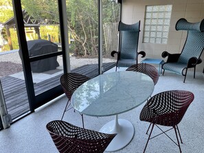 An outdoor patio with a glass table surrounded by wicker chairs, large windows, and a view of a wooden deck and trees. - Mid-Century Meets Boaters’ Dream (Siesta Key)