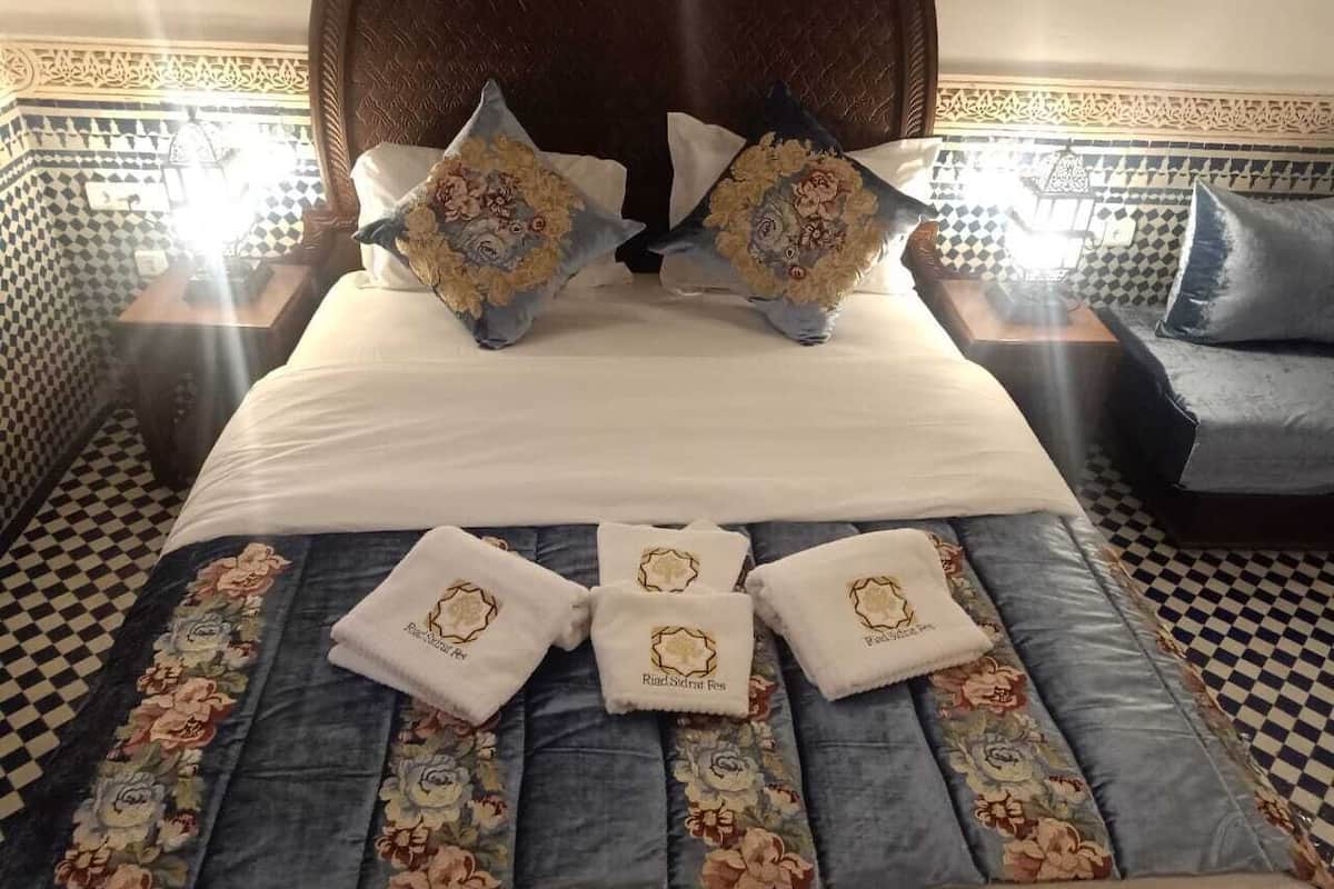 Cama bem arrumada com almofadas florais e toalhas combinando, chão xadrez e parede estampada.