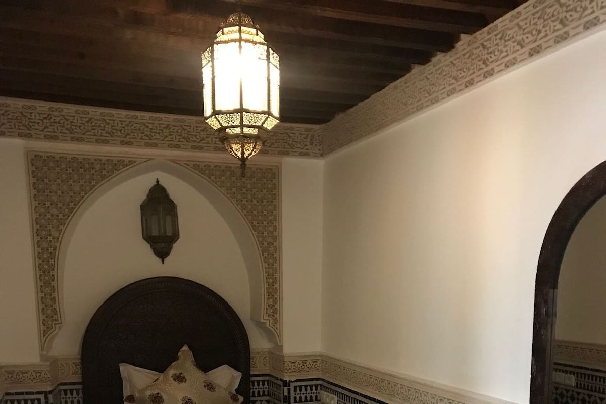 Quarto com uma cama, mesa de cabeceira de madeira, escrivaninha e espelho. O quarto possui piso de azulejos estampados e azulejos decorativos nas paredes.