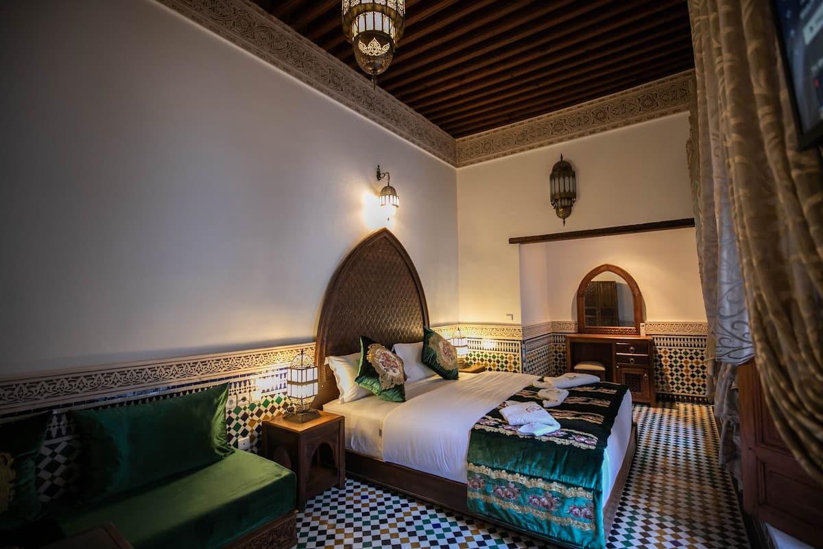 Quarto em estilo marroquino tradicional, com piso xadrez, cabeceira ornamentada e paredes estampadas.