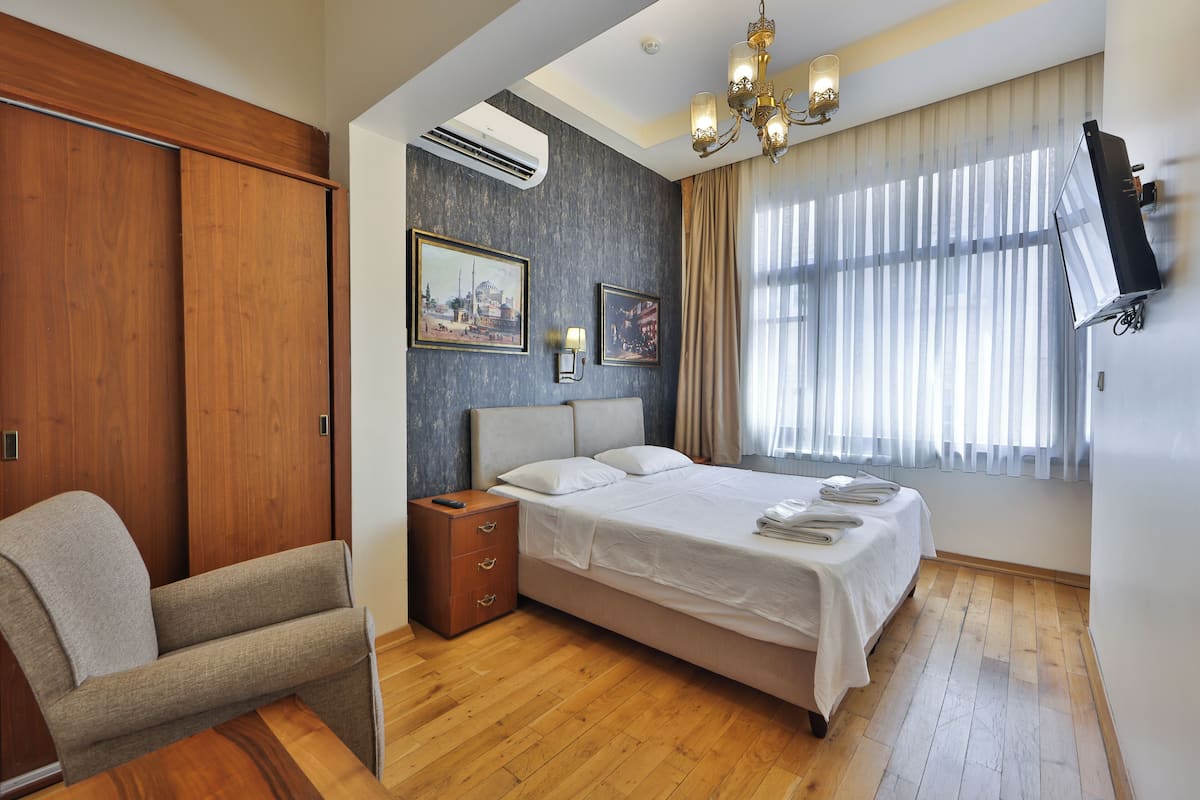Une chambre à coucher comprenant un lit, une table de chevet, une chaise, un téléviseur fixé au mur et une fenêtre avec des rideaux.