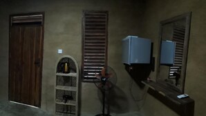 A room with a wooden door, a shelf with a kettle, a fan, and a wall-mounted TV.