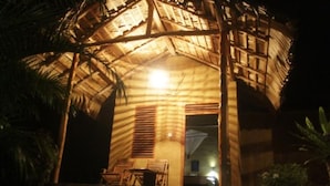 A thatched-roof structure with a porch and chairs, illuminated by a single light source.