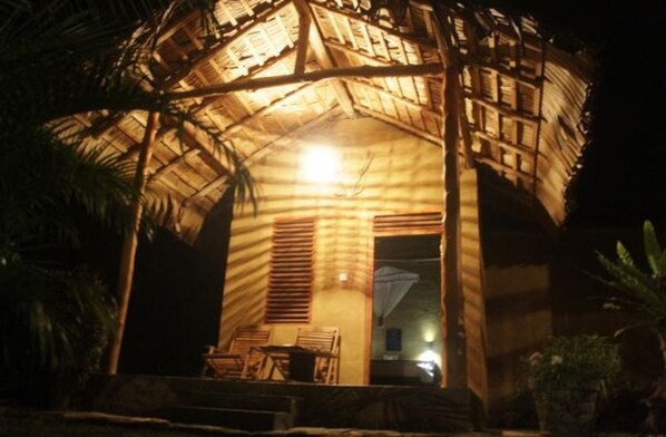 A thatched-roof structure with a porch and chairs, illuminated by a single light source. - Private Organic House (Udawalawa)