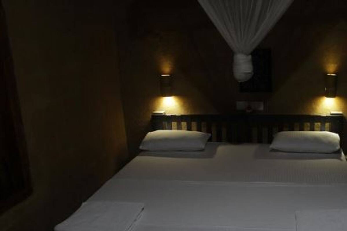 Une chambre d’hôtel à la lumière tamisée, avec deux lits, chacun doté d’un matelas et de coussins blancs. Deux lampes fixées au mur éclairent la pièce.