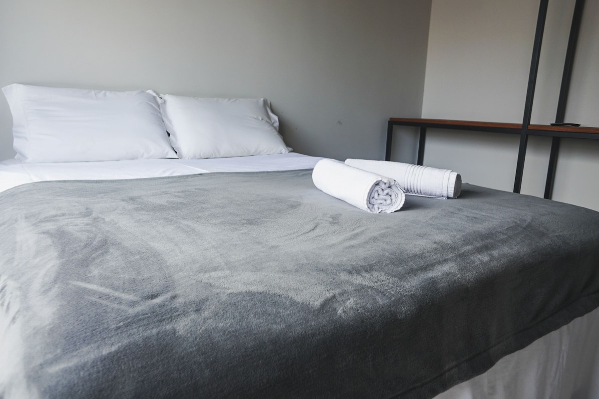 A neatly made bed with white linens and a grey blanket, a rolled white towel, and a wooden headboard.