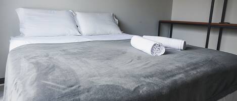 A neatly made bed with white linens and a grey blanket, a rolled white towel, and a wooden headboard.