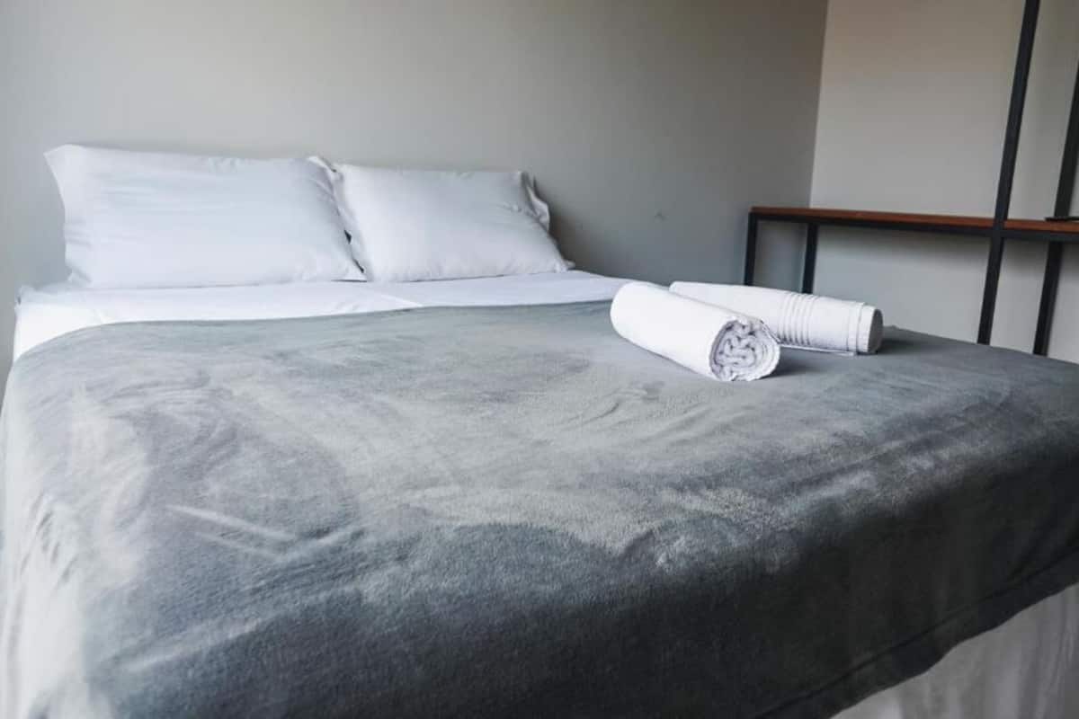 A neatly made bed with a grey bedspread and white pillows, a rolled white towel on the bed, and a wooden headboard.