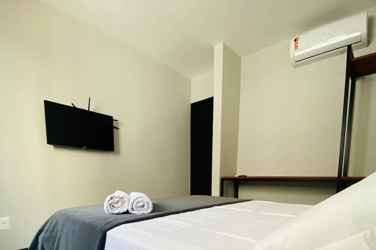 A hotel room with a bed, a flat-screen TV mounted on the wall, and an air conditioning unit.
