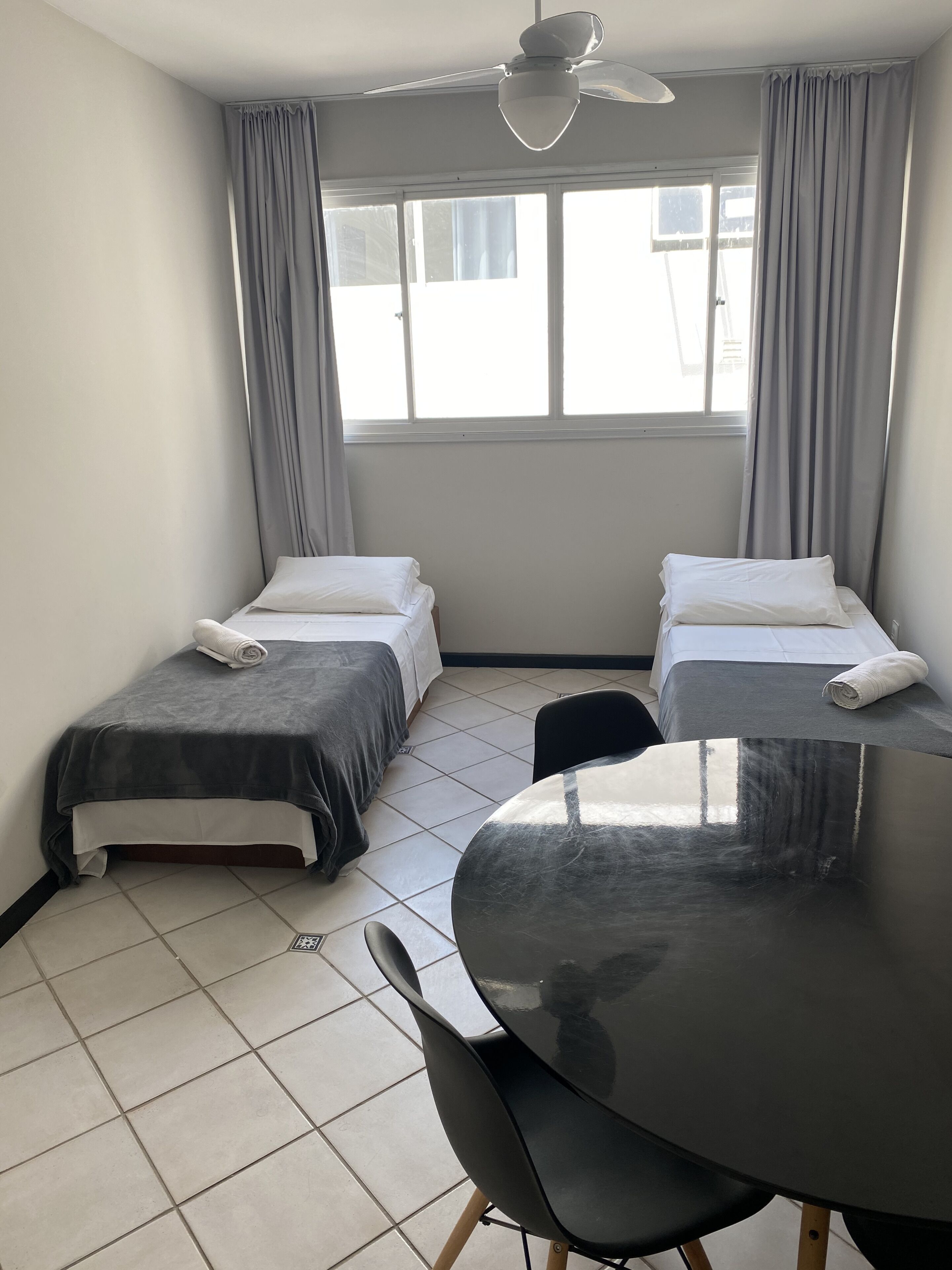 Una habitación pequeña y limpia con dos camas individuales, una mesa redonda y un ventilador de techo.