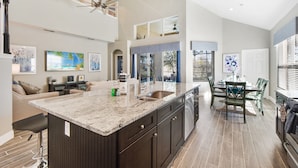 A modern kitchen with a large island, dark cabinetry, and a dining area with a table and chairs. - Ohana House: 5 Min to WDW! | 100% Renovated | Modern, Luxury, Lakeview, Gameroom (Kissimmee)