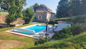 Une piscine entourée de chaises longues, un bâtiment en pierre et une végétation luxuriante.