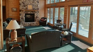 A cozy cabin interior with a stone fireplace, wicker furniture, and large windows offering views of the outdoors. - Round Lake Cabin Retreat (Hayward)