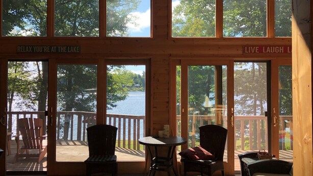 A cabin with large windows offering views of a lake and trees. There are two Adirondack chairs and a small table. Signs on the windows read "RELAX YOU'RE AT THE LAKE" and "LIVE LAUGH L." - Round Lake Cabin Retreat (Hayward)