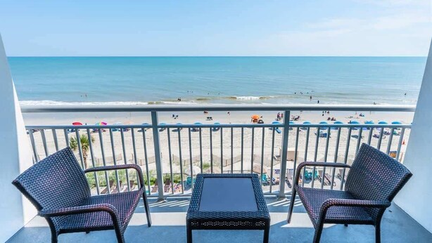 A balcony with two wicker chairs and a small table, overlooking a beach with people and umbrellas. - Remodeled 5th Floor Condo All Tile Near Boardwalk! (Myrtle Beach)