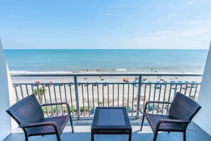 Ein Balkon mit zwei Korbstühlen und einem kleinen Tisch, mit Blick auf einen Strand mit Menschen und Sonnenschirmen.