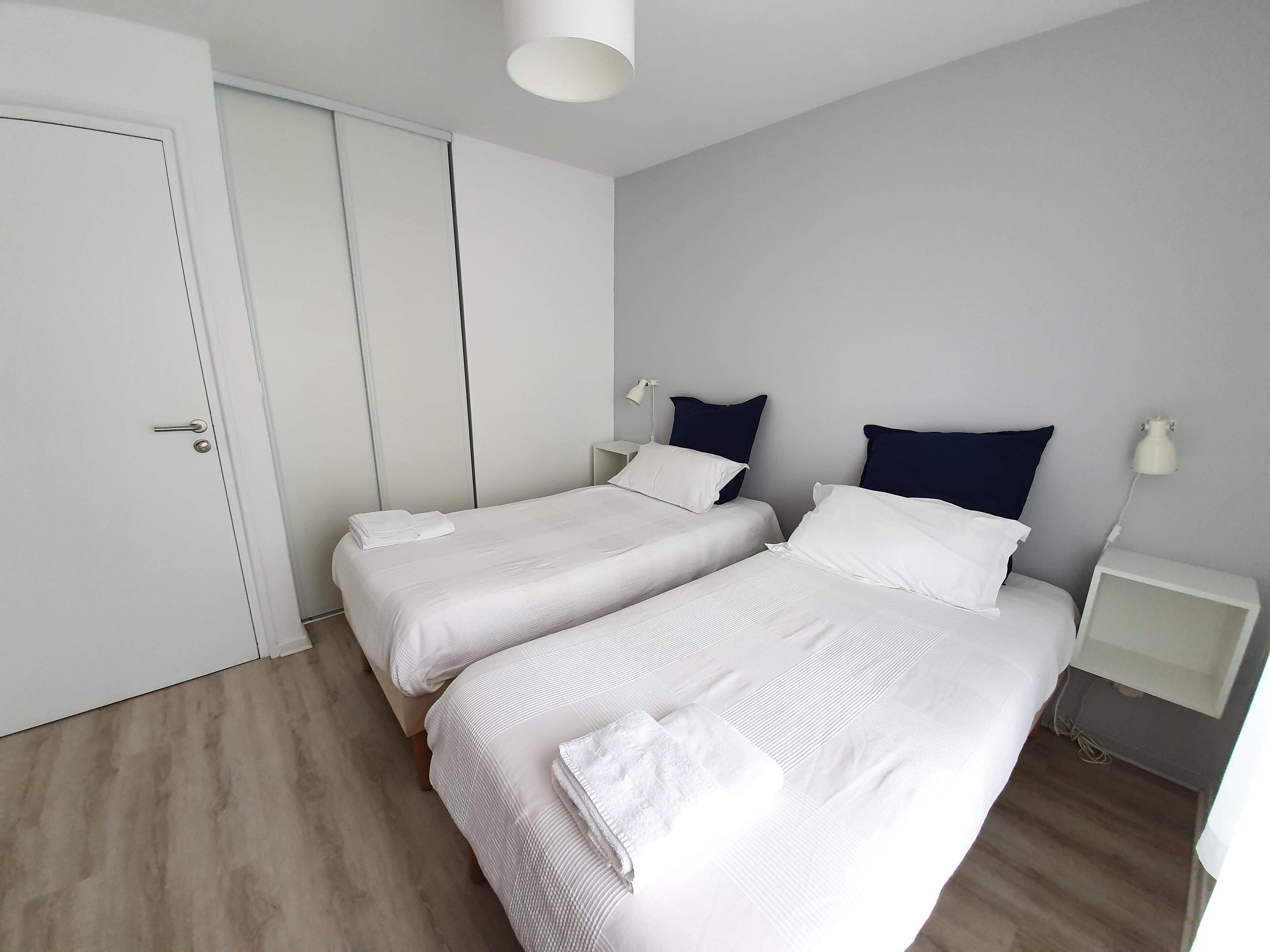 A modern hotel room with two single beds, a wardrobe, a bedside table with a lamp, and a small nightstand.