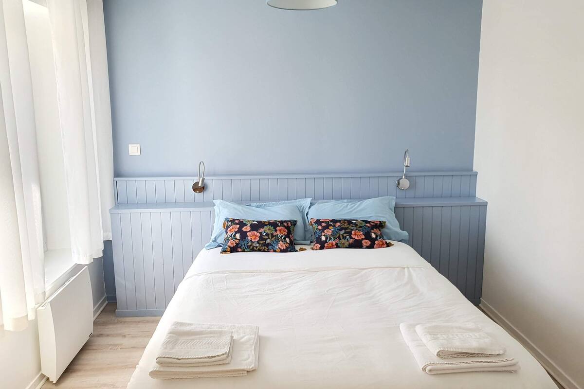 Quarto com cama, cortinas brancas, parede azul e luminária no teto.