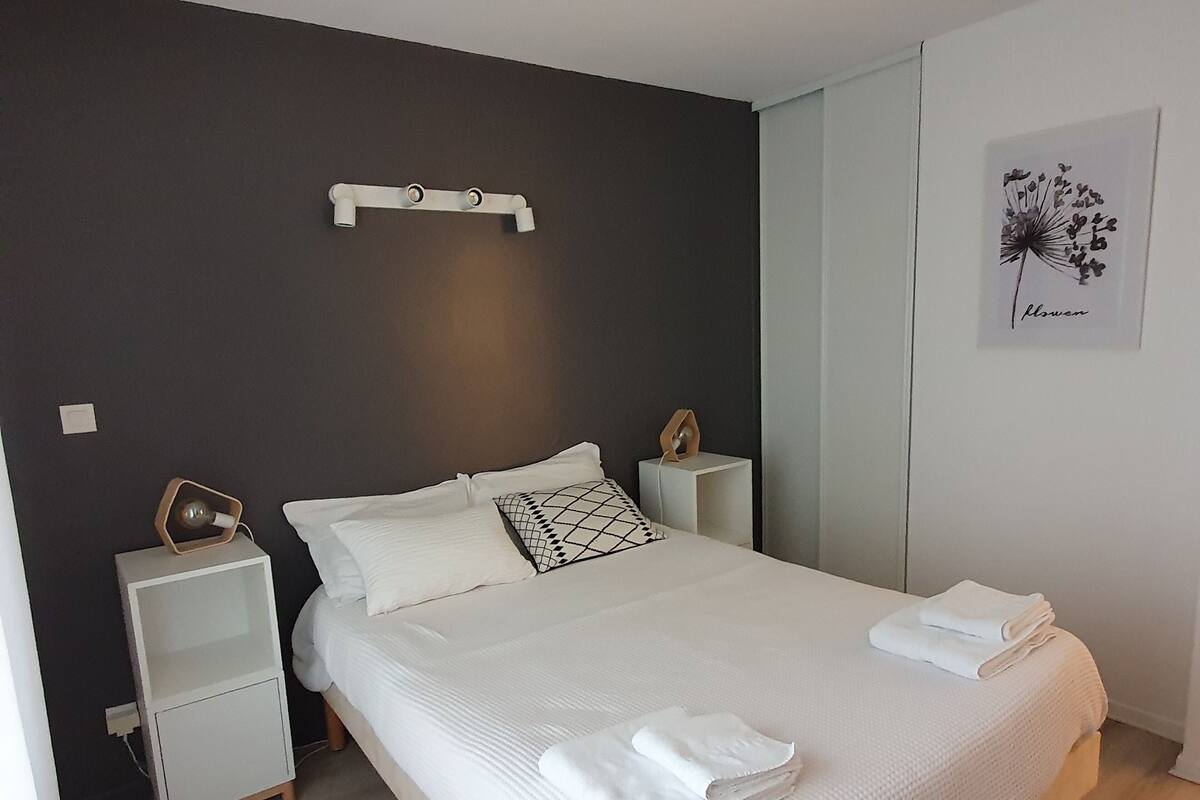 Uma cama bem arrumada com lençóis brancos e duas almofadas, uma mesa de cabeceira com abajur e uma prateleira na parede acima da cama.
