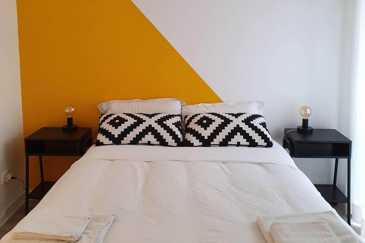 Quarto com cama, duas mesas de cabeceira, luminária pendente geométrica e uma parede em tom de amarelo.