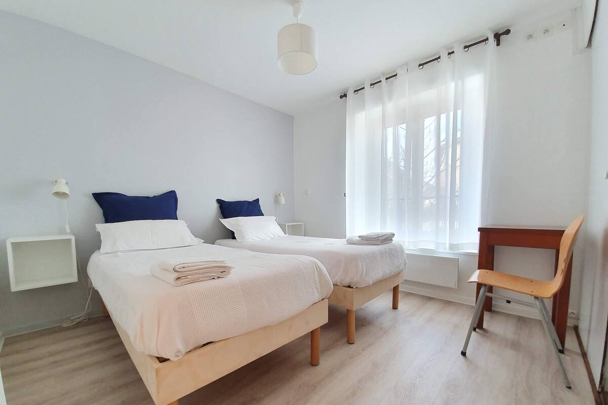 Quarto com duas camas, uma cadeira de madeira, uma mesinha e uma janela com cortinas.