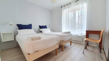 Quarto com duas camas, uma cadeira de madeira, uma mesinha e uma janela com cortinas.