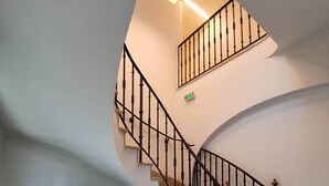 Een wenteltrap met zwarte spijlen en een witte muur. Een grote potplant staat aan de linkerkant, en een rieten mandje ligt op de vloer.