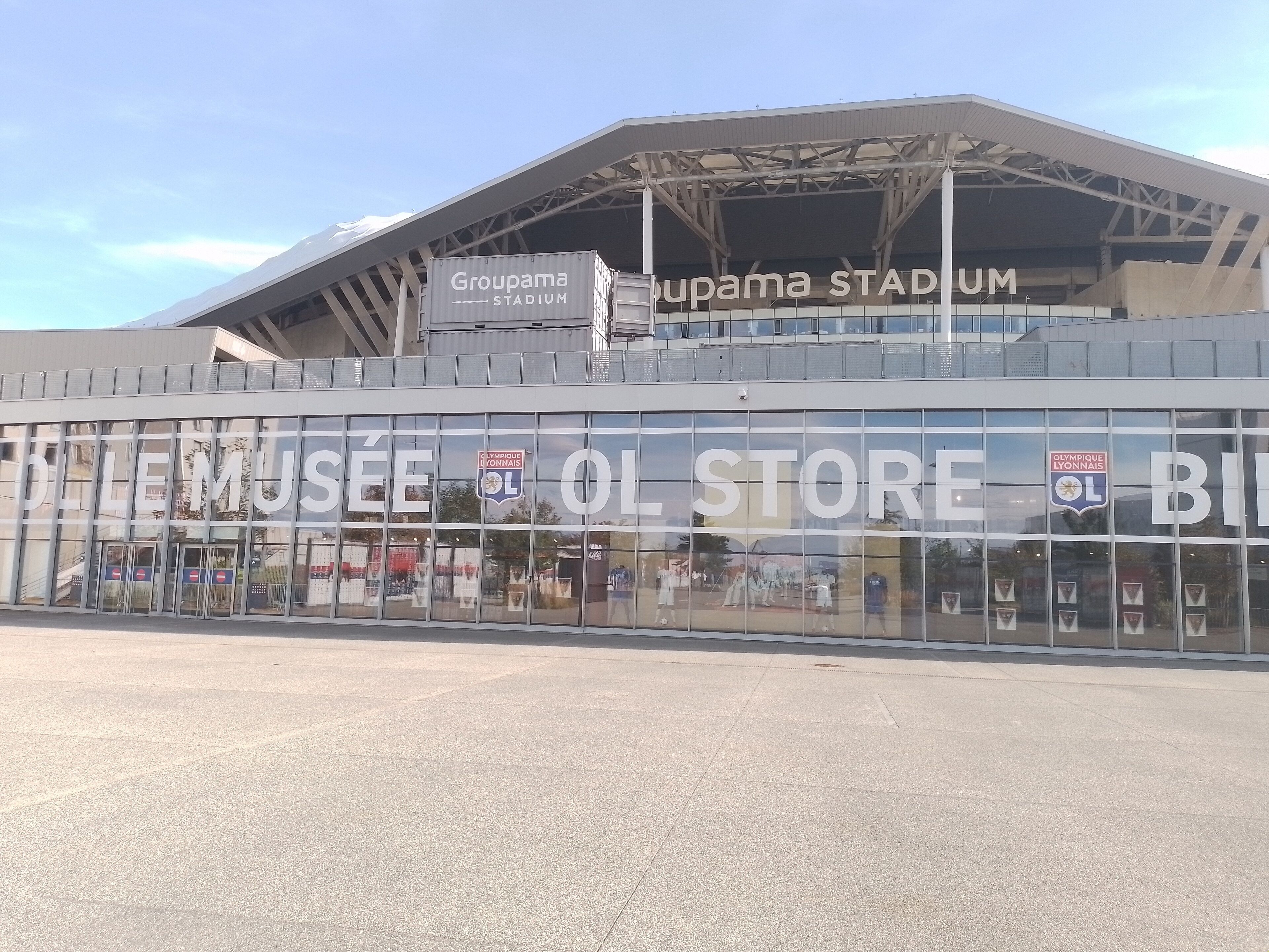 En stadion med en stor glasfasad och skyltar för Groupama Stadium och Olympique Lyonnais-butiken.
