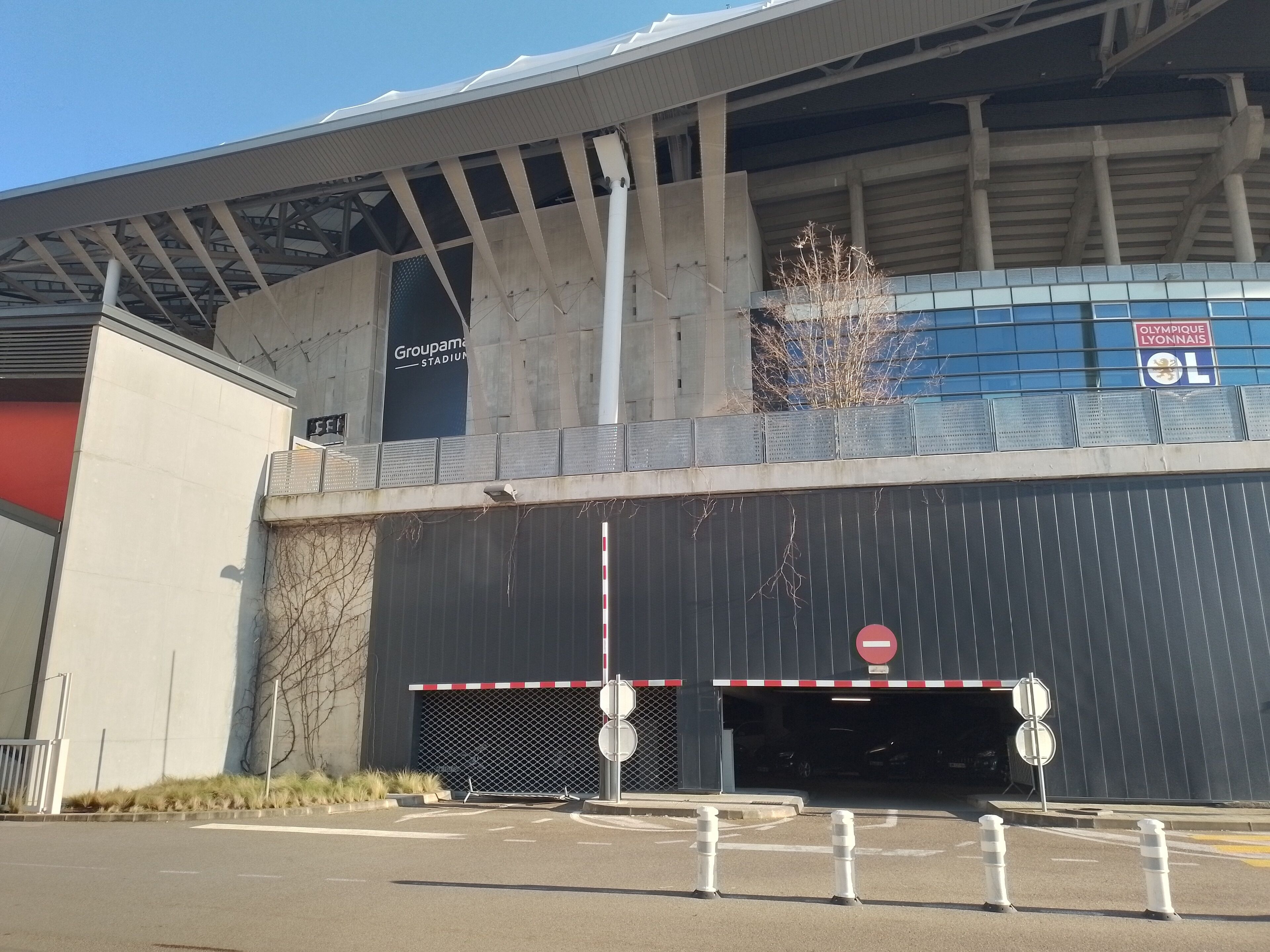 A stadium entrance with a no-entry sign, a Groupama Stadium sign, and a blue building with the Olympic Lyonnais logo.