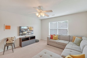 A living room with a sofa, a TV, a ceiling fan, and a window with blinds. - Family friendly w/ beautiful pool, SPA, and BBQ terrace! (Tomball)
